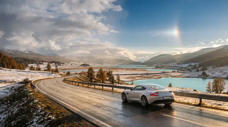 A silver touring car driving along a quiet winding road through snow-dusted alpine valleys and turquoise lakes under sunlit afternoon clouds.